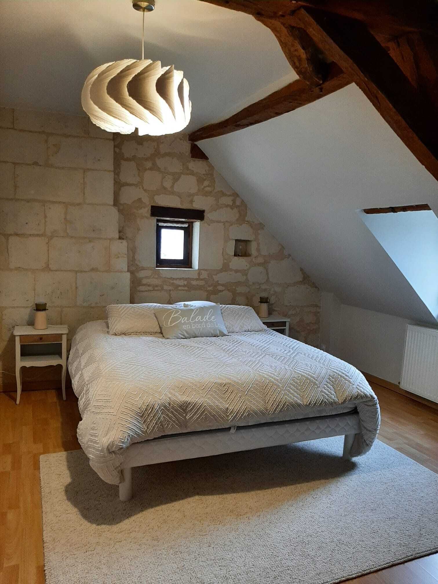 Een slaapkamer met een stenen muur, een schuine plafond, een bed met een beddegoed met patroon, een hanglamp en een klein raam.