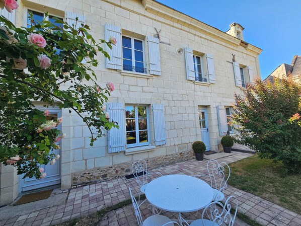 A two-story building with white stone walls and blue shutters. There is a white outdoor table and chairs, and a small garden area with a potted plant. - AU PIED DU LAVOIR (Gennes-Val-de-Loire)