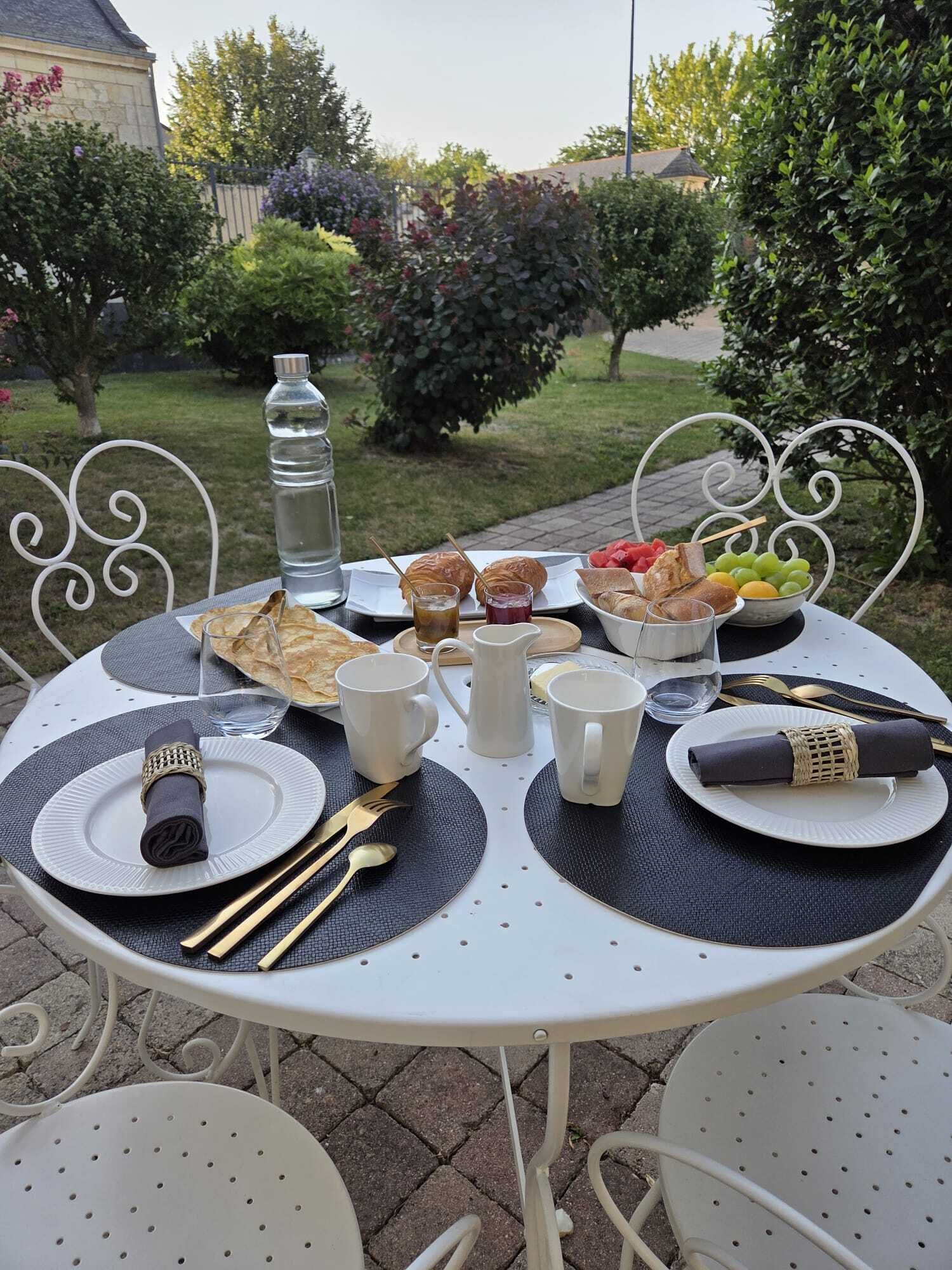 Un coin repas aménagé à l’extérieur, avec une table dressée pour deux personnes, comprenant une bouteille d’eau, du pain, des fruits et une carafe.