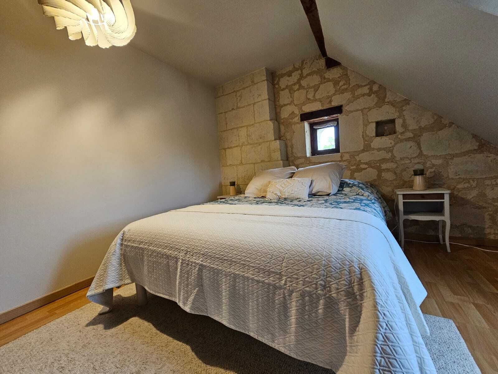 A bedroom with a stone wall, a bed with a quilted cover, a nightstand, and a small window.