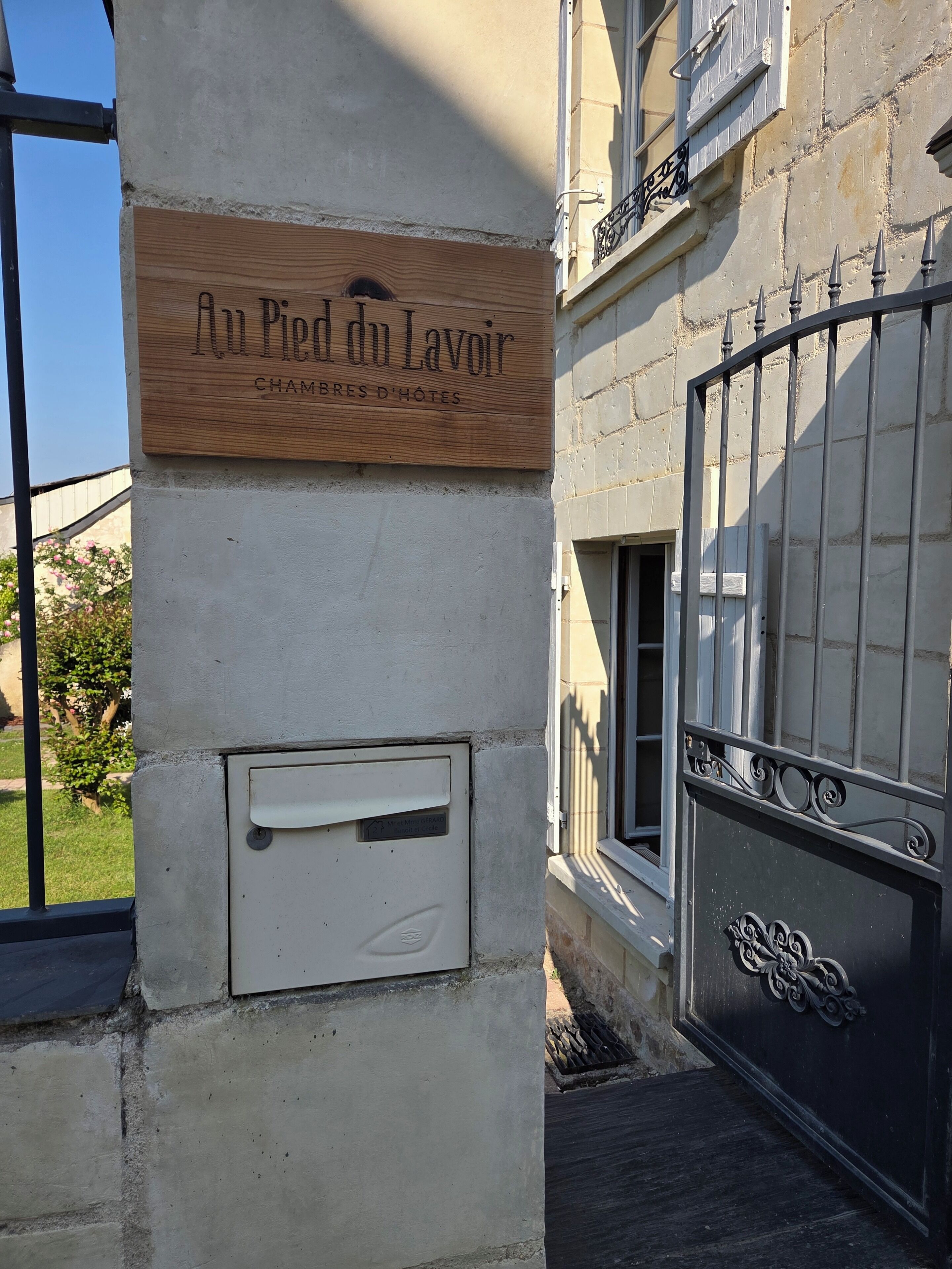 A wooden sign with "Au Pied du Lavoir" and "CHAMBRES D'HOTES" indicating a hotel. A white mailbox is attached to the wall.