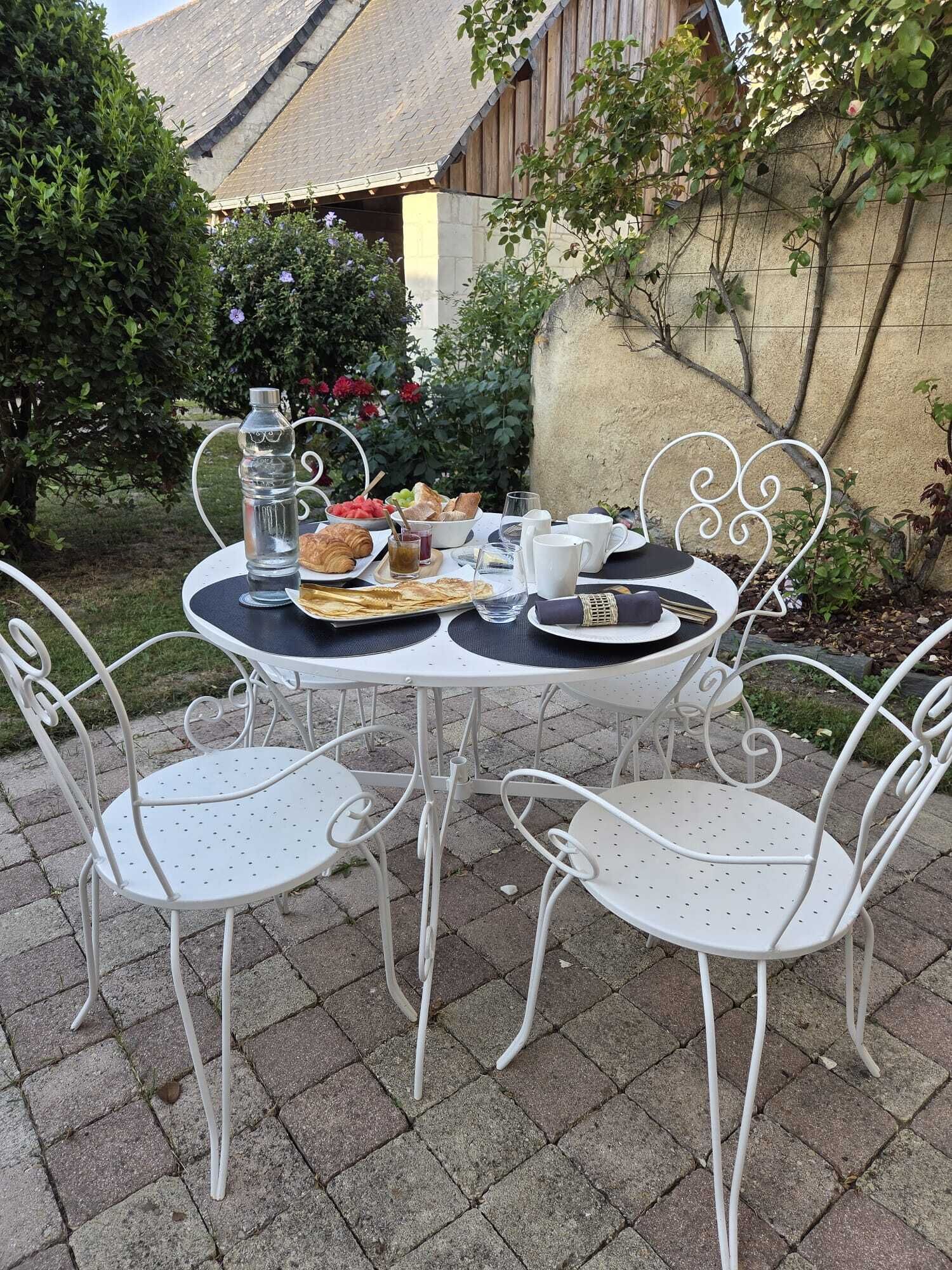 Taula de pati amb caires de metall blanc, assortiment de menjar matinal amb croissants, fruita i begudes, i un mur de pedra al fons.