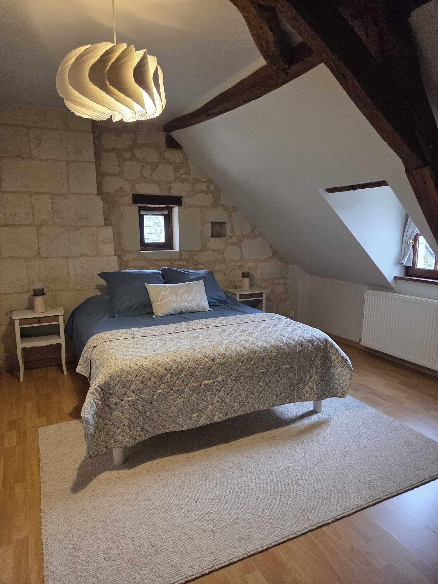 Un dormitorio con pared de piedra, techo inclinado, cama con colcha estampada, mesita de noche, lámpara y lámpara colgante.