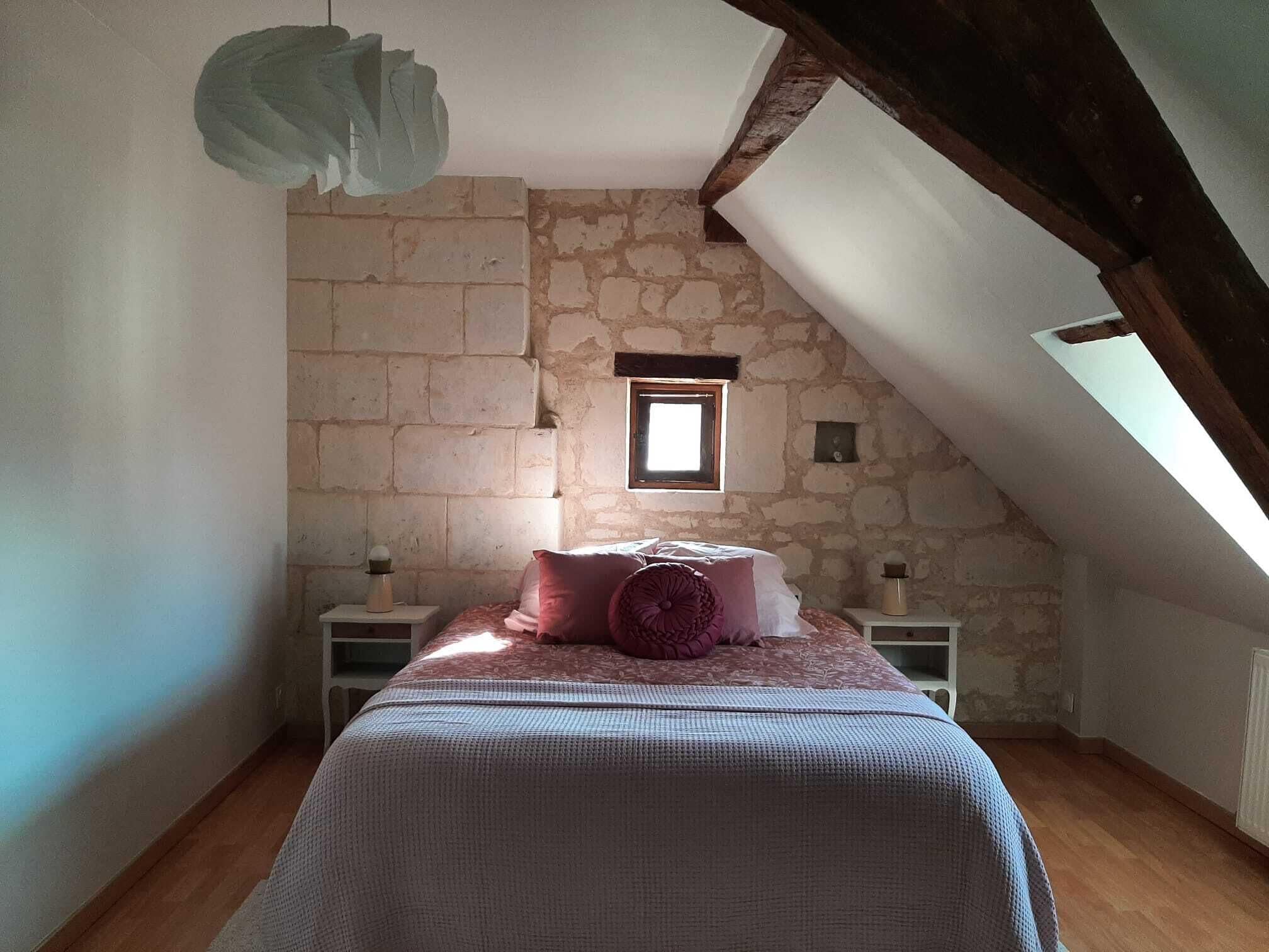 Un dormitorio con techo inclinado, paredes de piedra, una ventana pequeña, una cama con colcha estampada, dos mesitas de noche con lámparas, y una lámpara colgante.