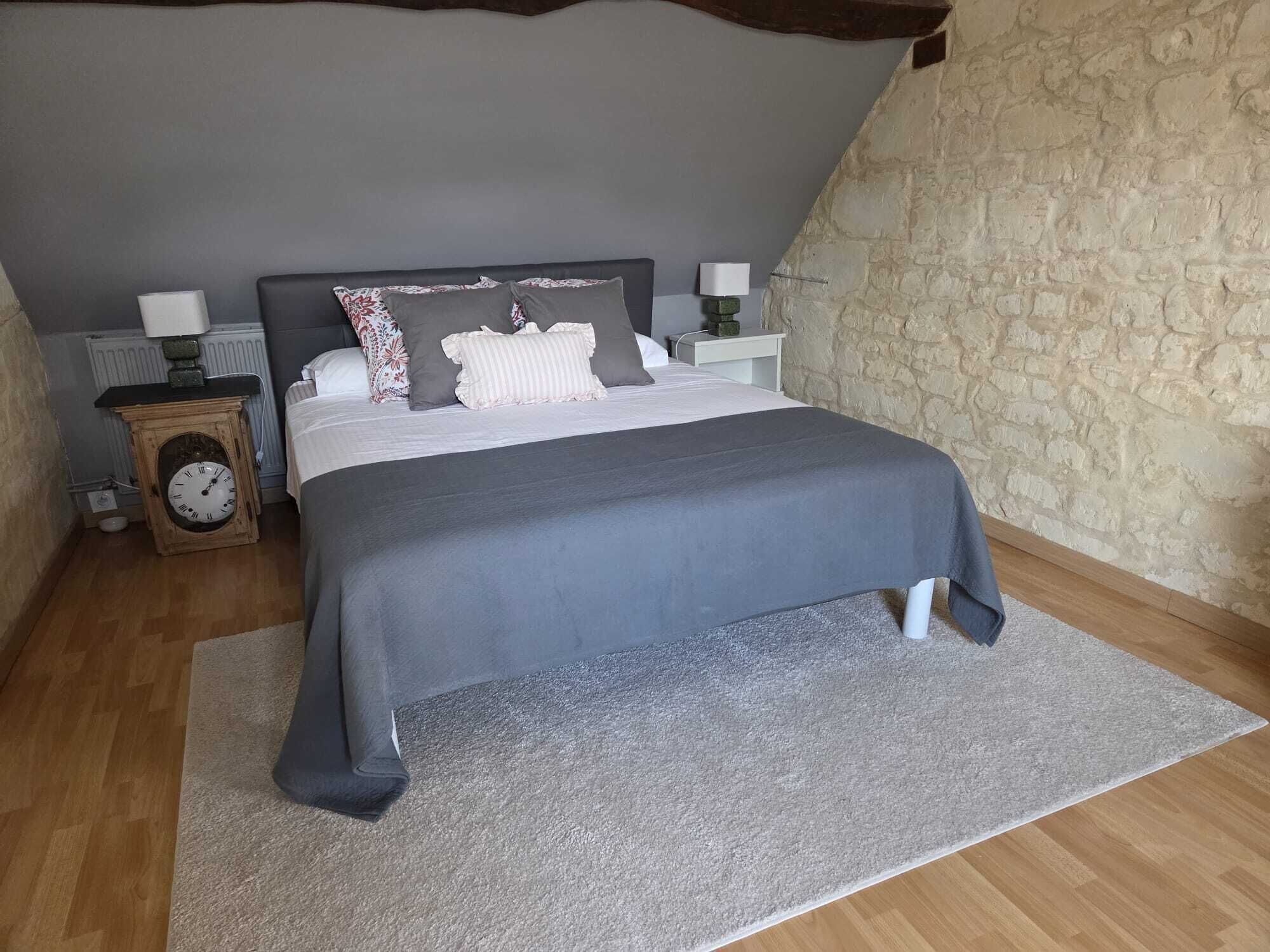 Un dormitorio con cama, un reloj, una mesita de noche y una pared de piedra.