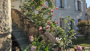 Ένα κτίριο από πέτρα με λευκά παντζούρια και ένας κήπος γεμάτος με ανθισμένες ροζ τριανταφυλλιές.