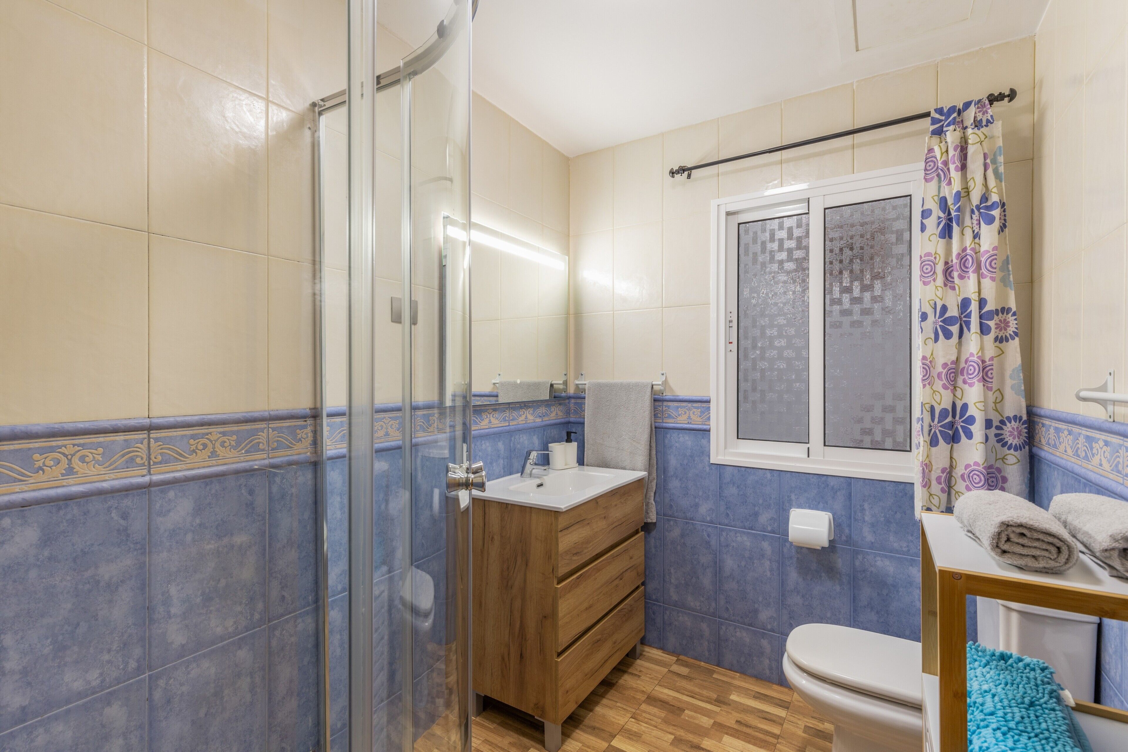 Ein Badezimmer mit Duschkabine aus Glas, einem Holzwaschtisch, einer Toilette und einem Fenster mit gemustertem Vorhang.