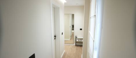 A narrow hallway with white walls and wooden flooring, leading to a room with a bed and a window.