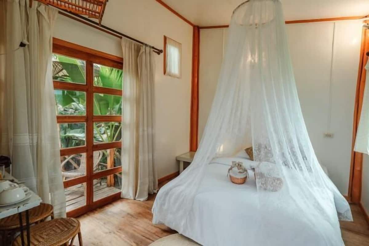 A bedroom with a bed under a canopy, a window with curtains, a small table, and a chair.
