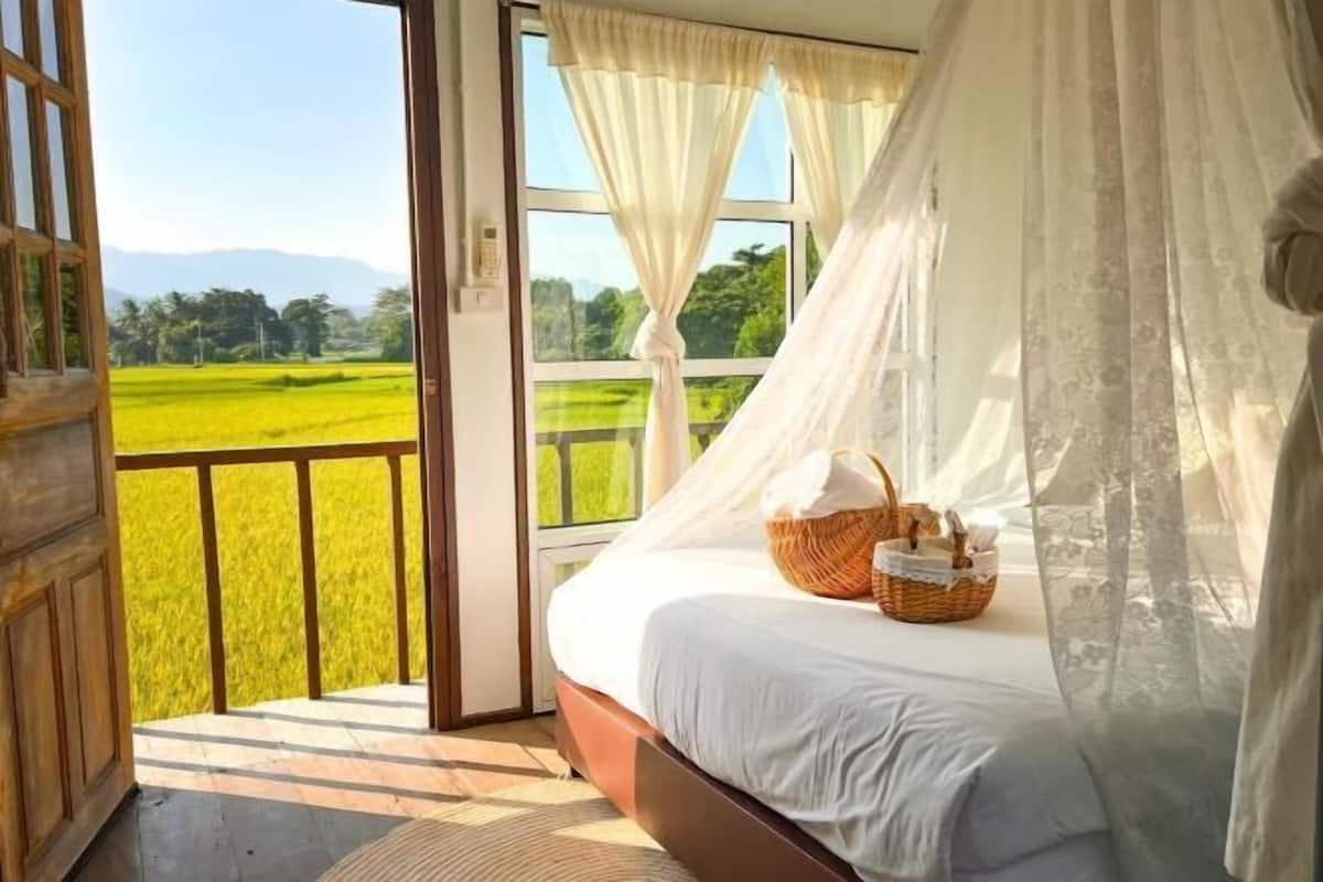 A bedroom with a large window, a bed with white linens, a wicker basket, and a view of a green landscape.