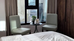 Une chambre d’hôtel avec un grand lit, deux fauteuils, une petite table et une vue sur la ville grâce à de grandes fenêtres.