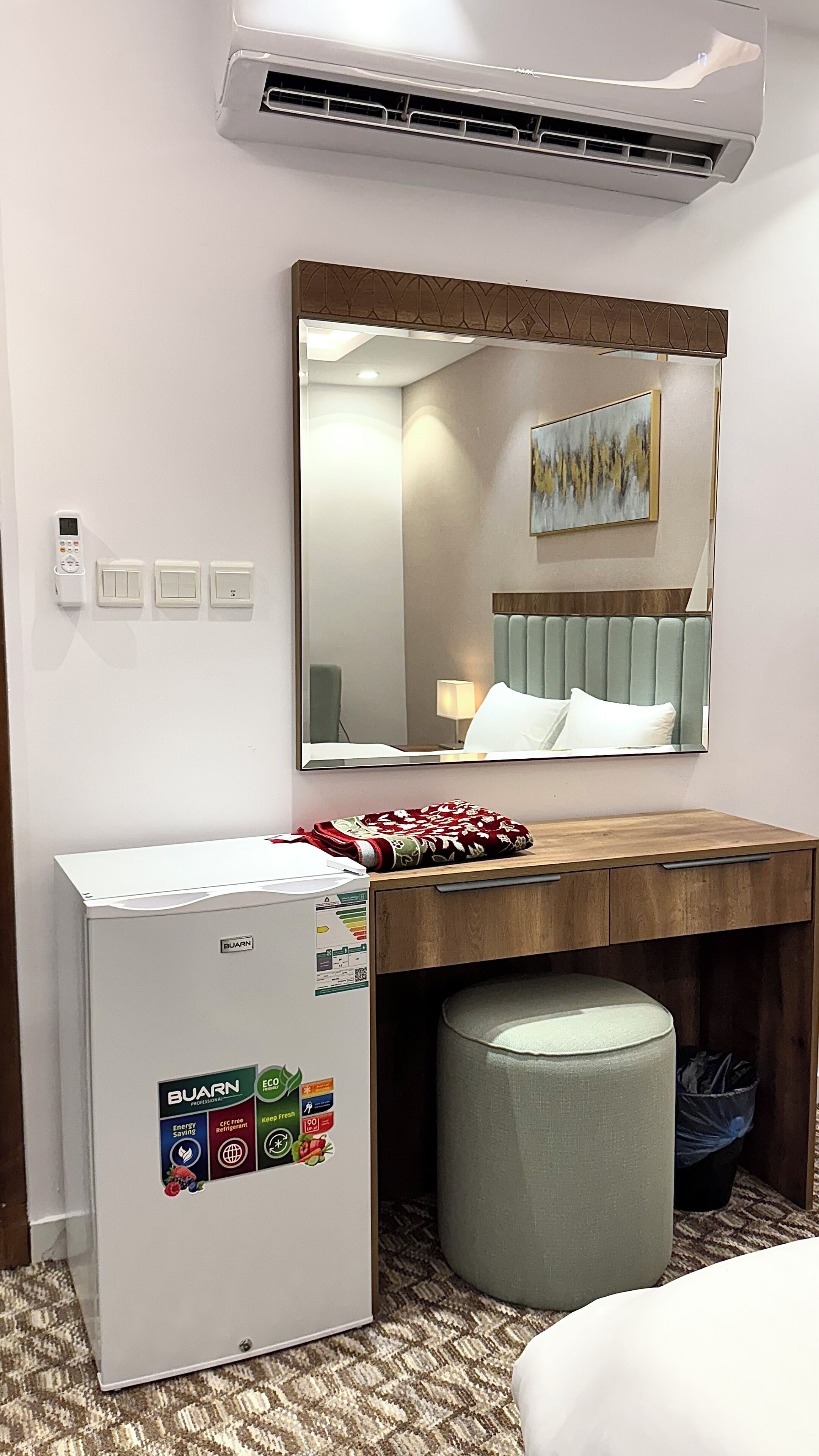 Quarto de hotel com cama, escrivaninha com computador, ar condicionado fixado na parede, espelho e termostato na parede.