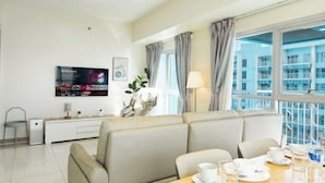 Une chambre d’hôtel moderne, dotée d’un canapé, d’une table à manger avec assiettes et verres, d’un téléviseur fixé au mur et offrant une vue sur la ville grâce à de grandes fenêtres.
