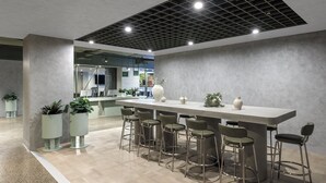 Un área de bar moderna con una barra larga y varios taburetes, rodeada de plantas en macetas y un espejo grande.