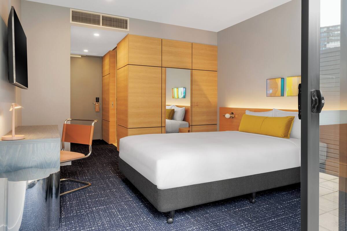 A hotel room with a large bed, a desk with a chair, a wardrobe, a TV, and artwork on the walls.