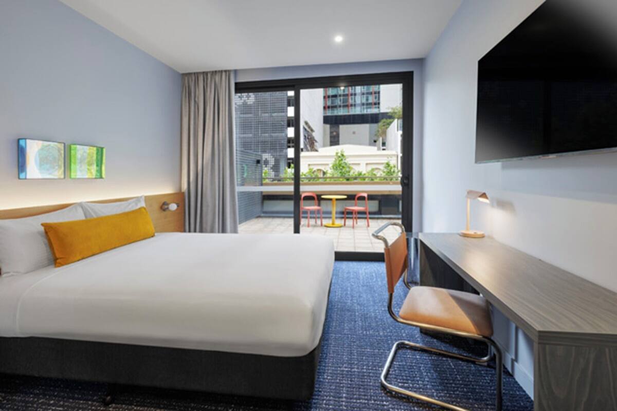 A hotel room with a bed, a desk, a chair, a TV, and a view of the outside.