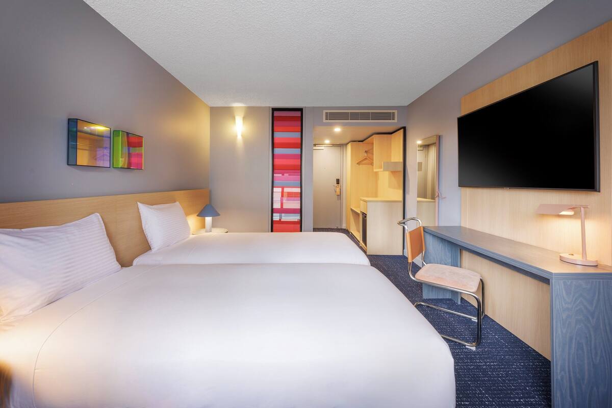 A hotel room with two beds, a desk, a chair, a TV, and a bathroom visible through an open door.