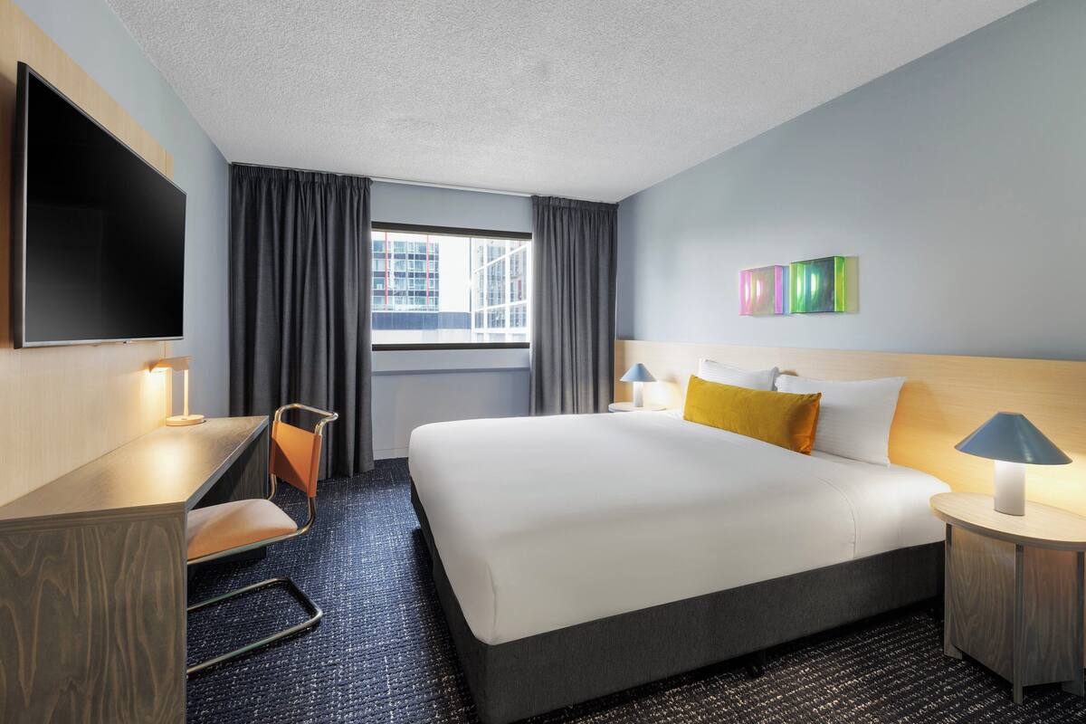 A hotel room with a large bed, a desk with a chair, a TV, and a window with curtains.
