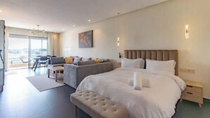 A modern hotel room with a large bed, a sofa, a small table, and a view of the city through large windows. - Horizon Marina terrace (Casablanca)