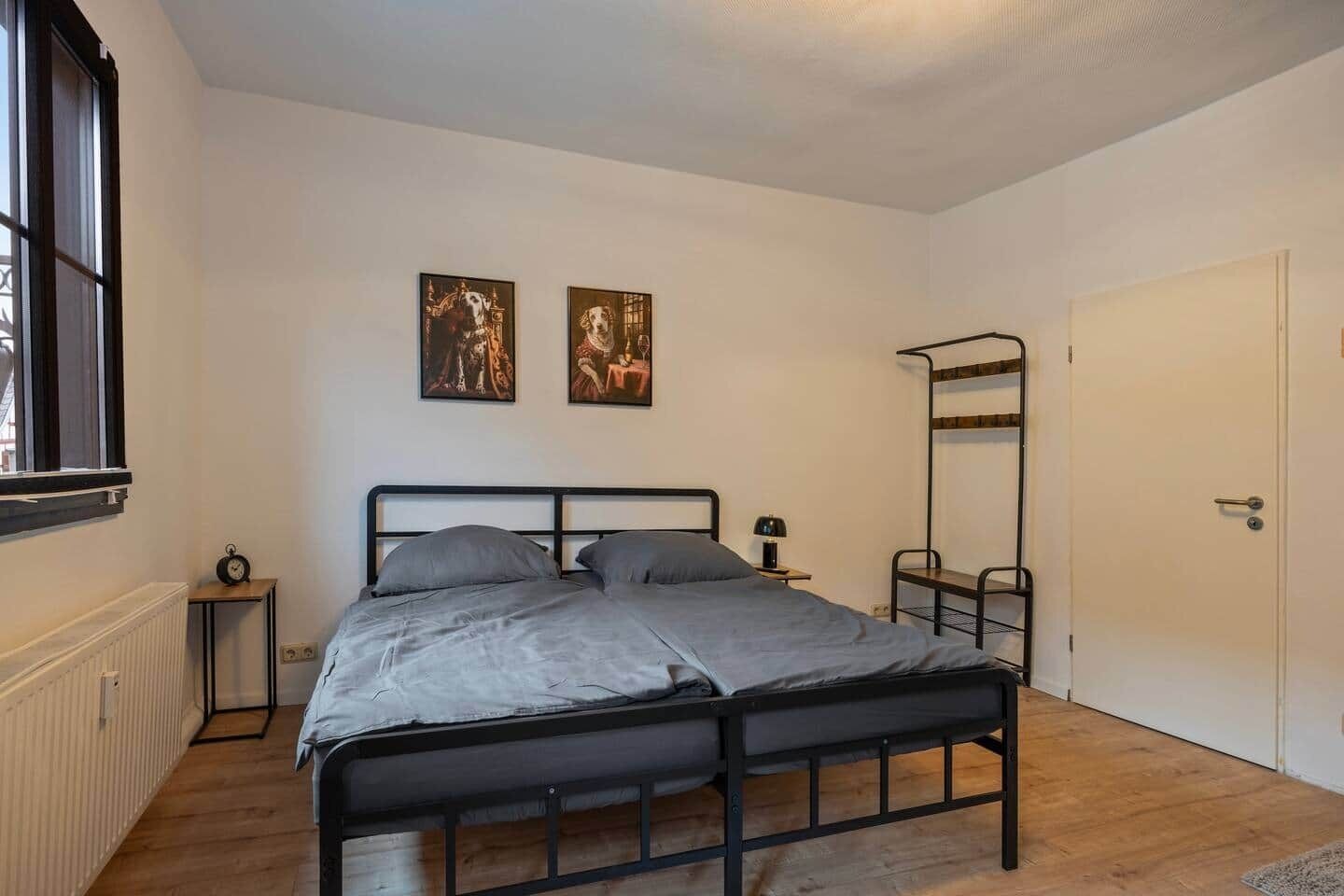 Een slaapkamer met een metalen bedframe, een nachtkastje met een lamp, een radiator en een raam met zonwering.