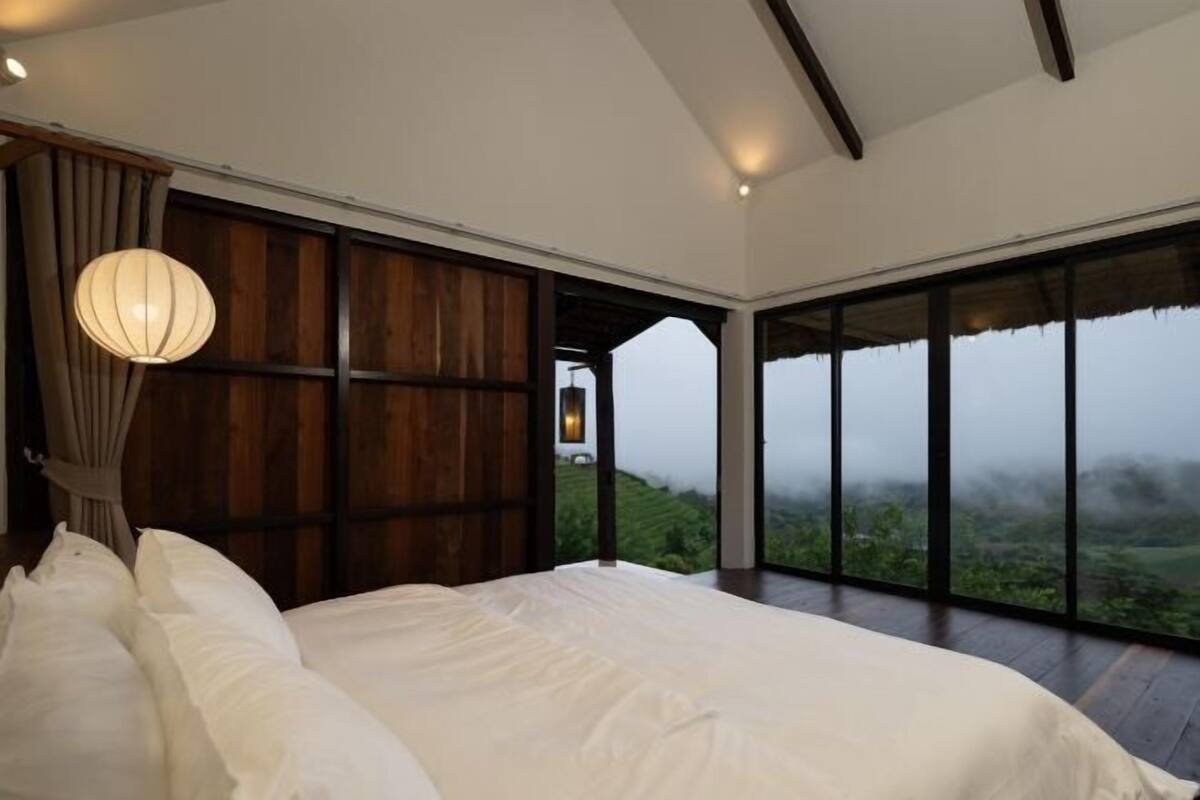 A bedroom with a large bed, a hanging light fixture, and a view of a misty landscape through large windows.