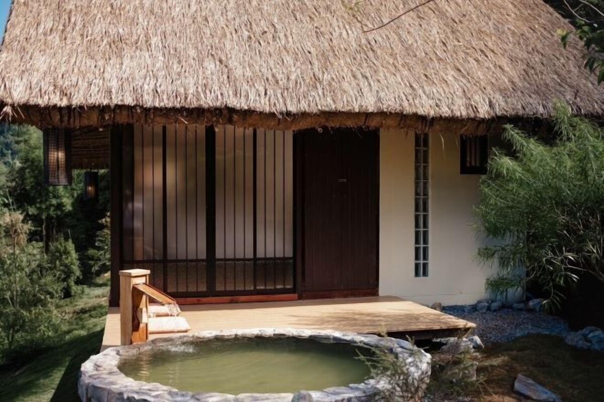 A traditional thatched-roof building with a wooden deck and a stone hot tub.
