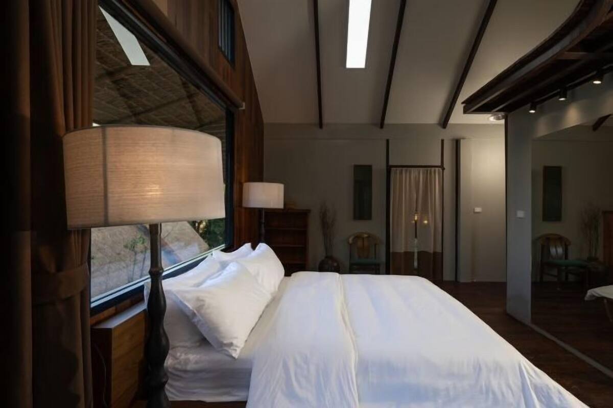A bedroom with a large bed, a bedside lamp, and a view of the outdoors through a window.