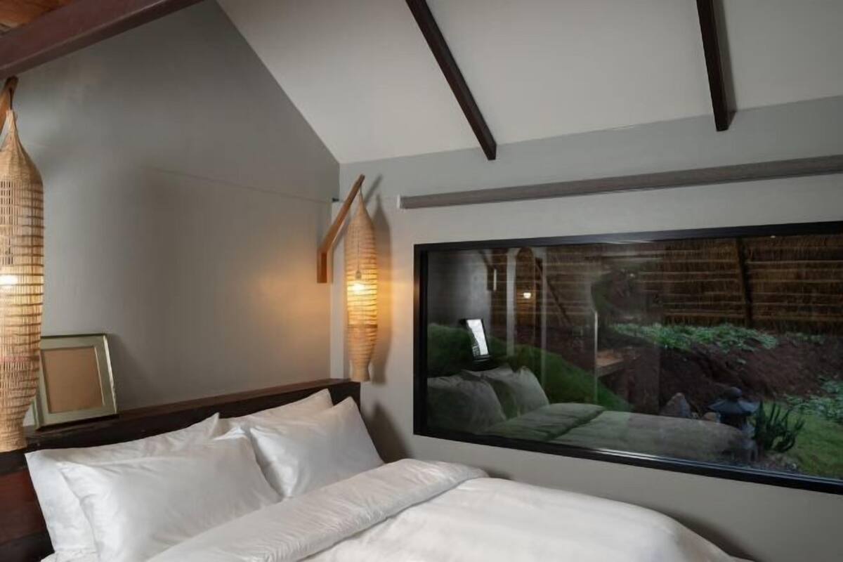 A bedroom with a large bed, a wooden ceiling, a wall-mounted TV, and a view of a garden through a window.