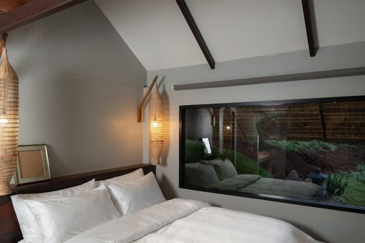 A bedroom with a large window, a bed with white linens, and a wooden ceiling.