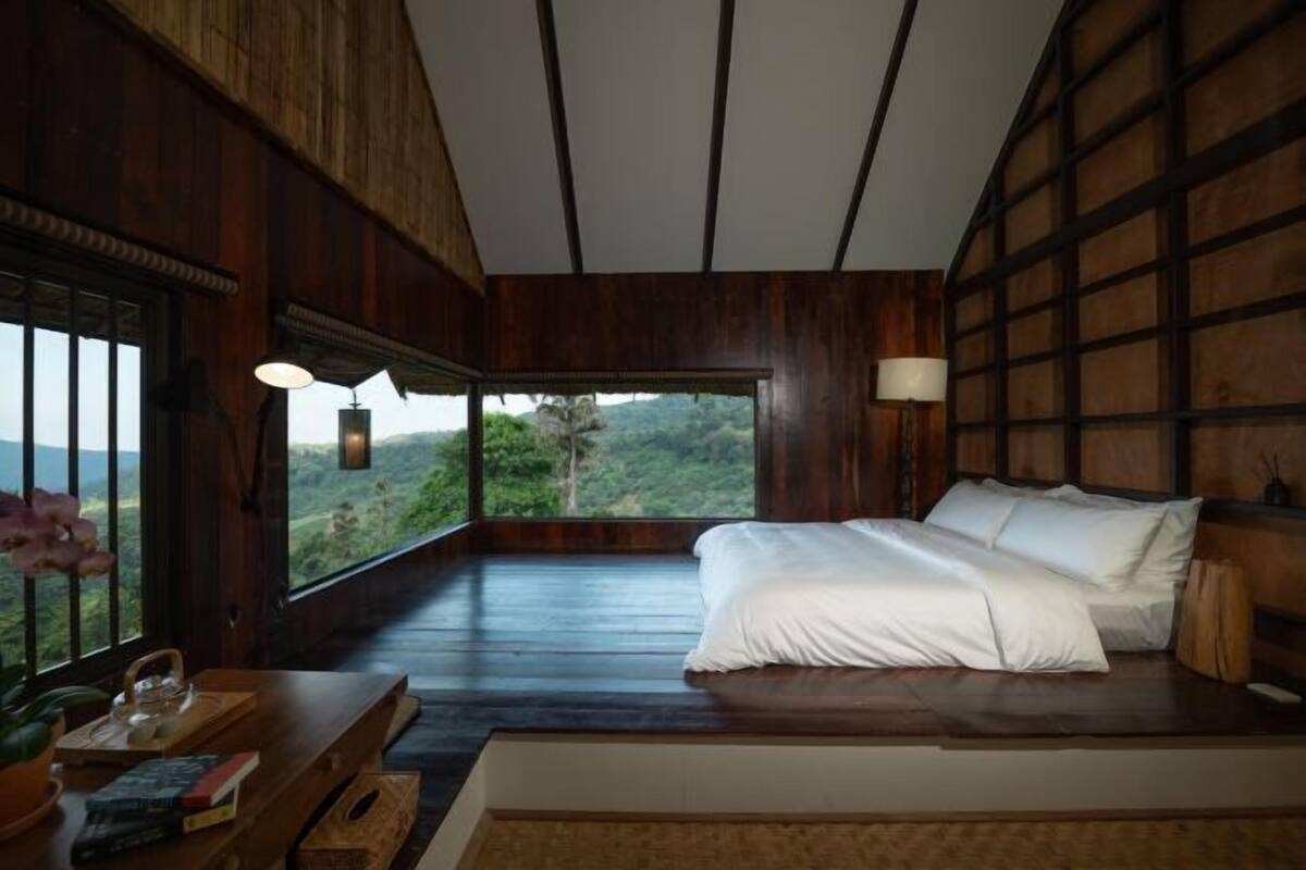 A bedroom with a bed, a nightstand, a window with a view, and a wooden ceiling.