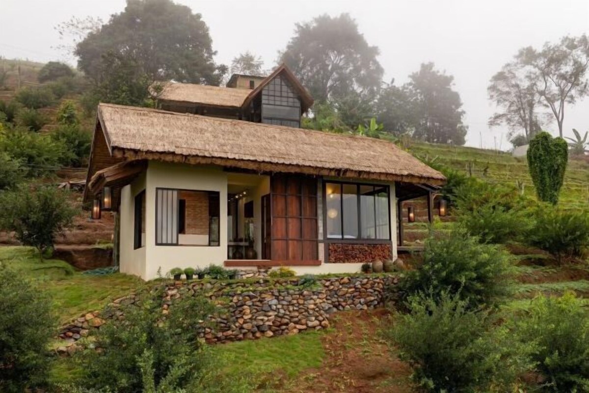 A small, single-story house with a thatched roof, surrounded by greenery and a stone wall.