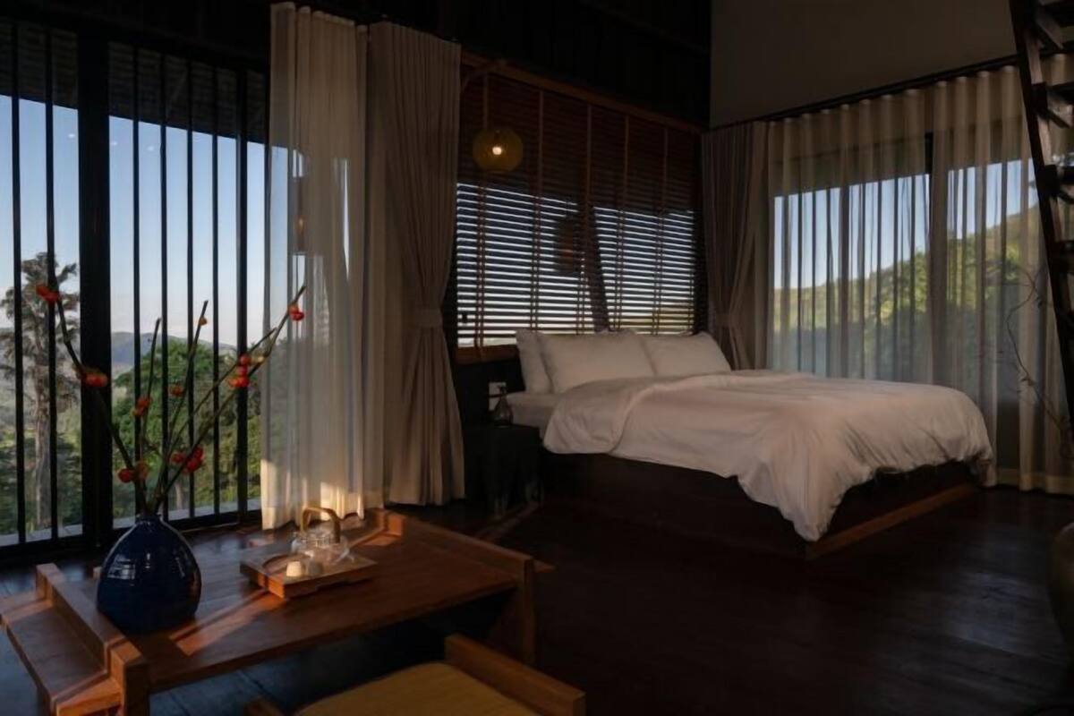 A bedroom with a bed, a bedside table, a vase with flowers, and a view through large windows.