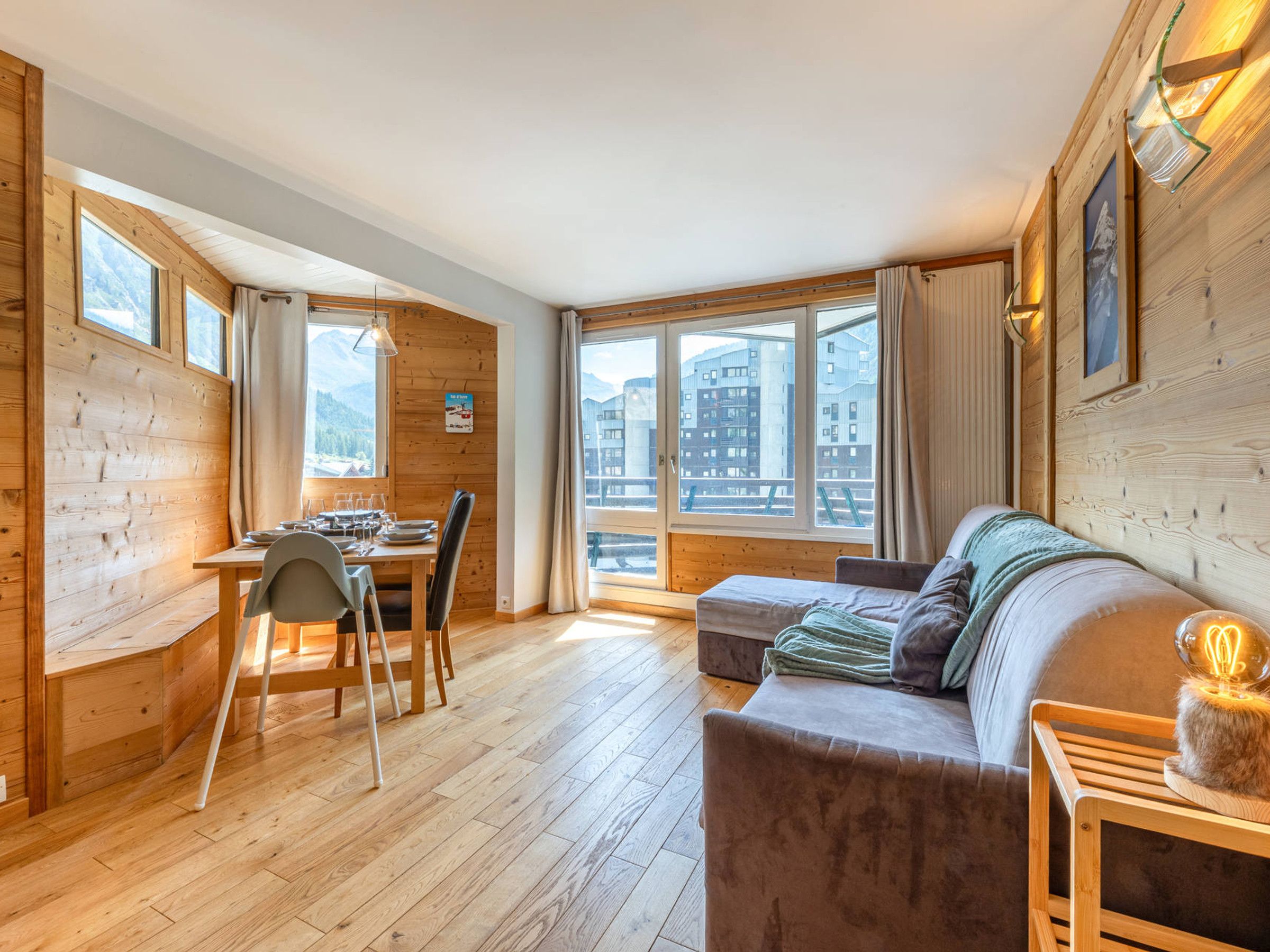 A cozy room with wooden walls, a dining area with a table and chairs, a sofa, and large windows offering a view of buildings and mountains.