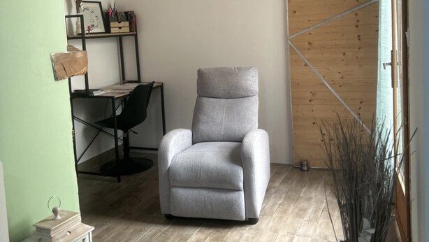 A room with a wooden ceiling, a grey armchair, a desk with a chair, a white wall, and a wooden door. - T2 in charming Ardèchois village (SAINT-JULIEN-EN-SAINT-ALBAN)