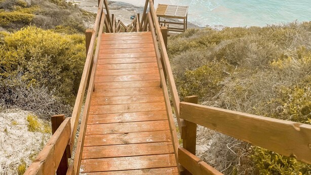Une promenade en bois qui mène à une plage aux eaux turquoise et limpides et au rivage sablonneux.