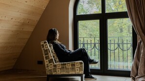 Ένα άτομο κάθεται σε μια καρέκλα με σχέδια σε ένα δωμάτιο με λοξή ξύλινη οροφή και ένα μεγάλο, αψιδωτό παράθυρο.