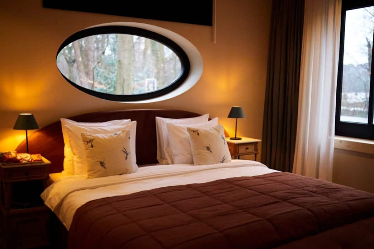 Une chambre d’hôtel avec un grand lit, deux tables de chevet avec des lampes, un grand miroir rond au-dessus du lit et une fenêtre avec des rideaux.