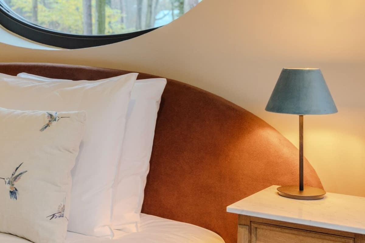Une chambre à coucher avec une fenêtre ronde, une tête de lit en bois, une table de chevet avec une lampe et des coussins à motifs floraux.