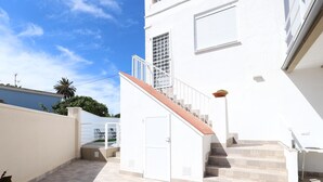 Un edificio moderno con una escalera que lleva a una puerta, un espacio cubierto para sentarse al aire libre y un cielo azul claro.