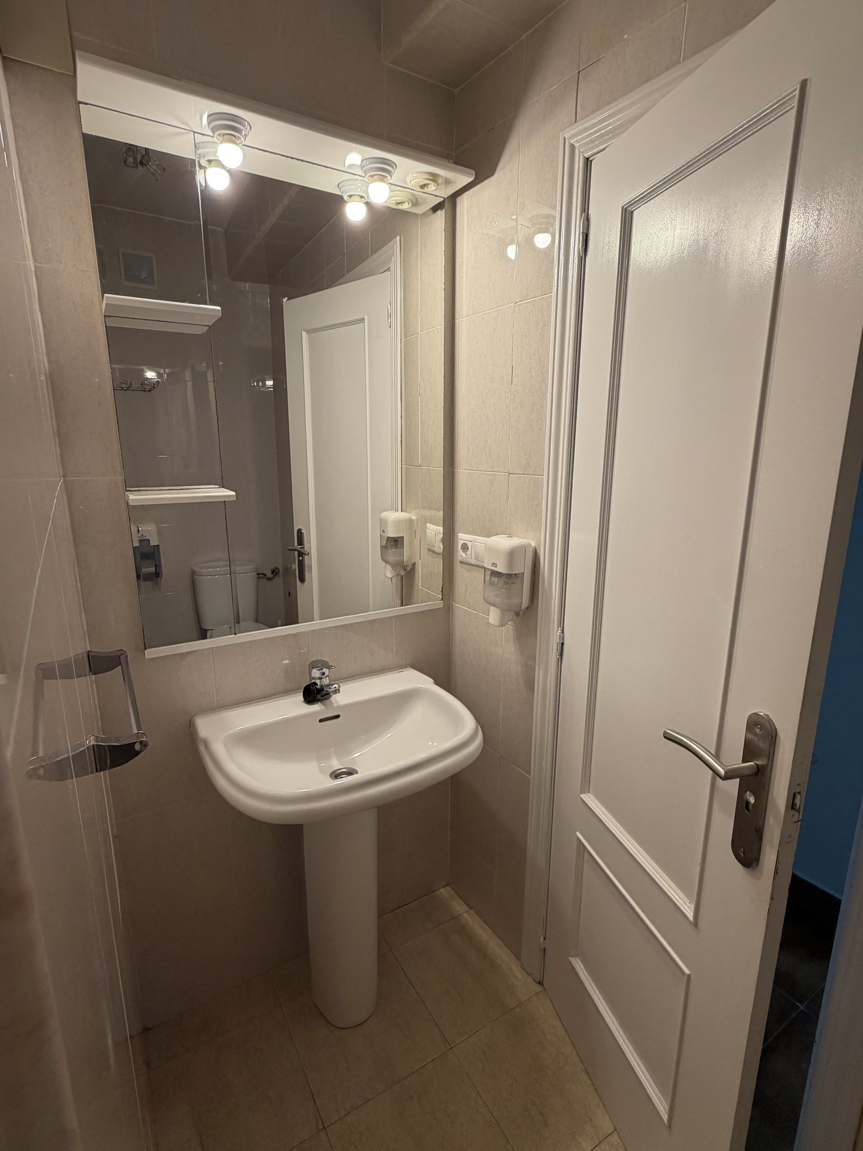 A bathroom with a pedestal sink, a mirror with lights, a door, and a hand dryer.