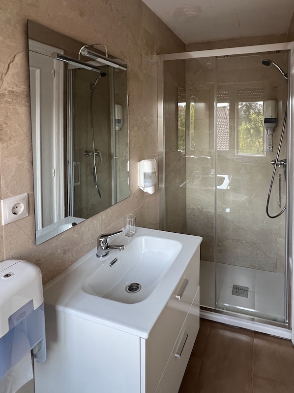 A modern bathroom with a large mirror, a white sink, a handheld showerhead, and a glass-enclosed shower. - ENTRE PUERTAS (Aranda de Duero)