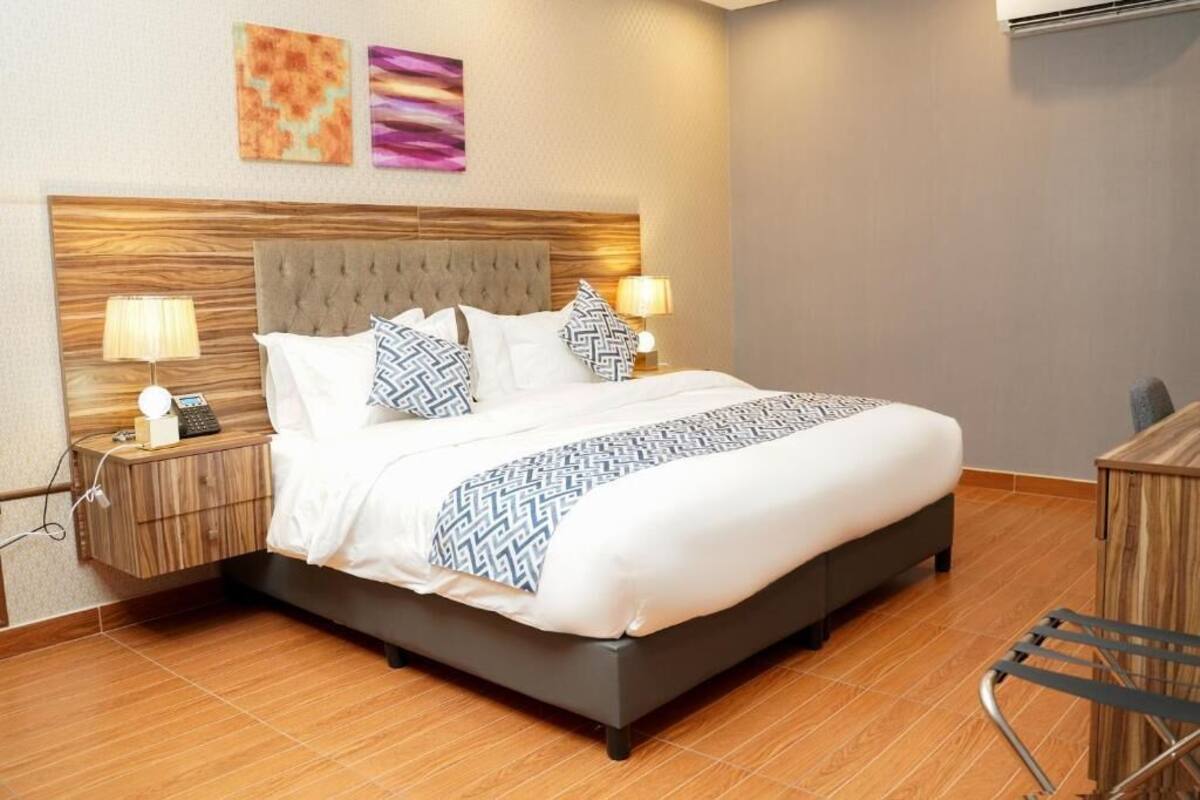 Une chambre d’hôtel moderne avec un grand lit, une tête de lit en bois, deux lampes de chevet et des œuvres d’art accrochées au mur.