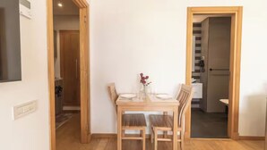 A dining area with a wooden table and chairs, a vase with flowers, and a view into a kitchen and bathroom through an open door.