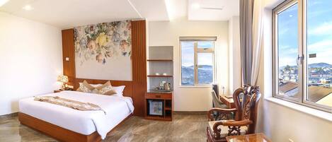 A hotel room with a large bed, a desk, a chair, a small refrigerator, and a window with a city view.