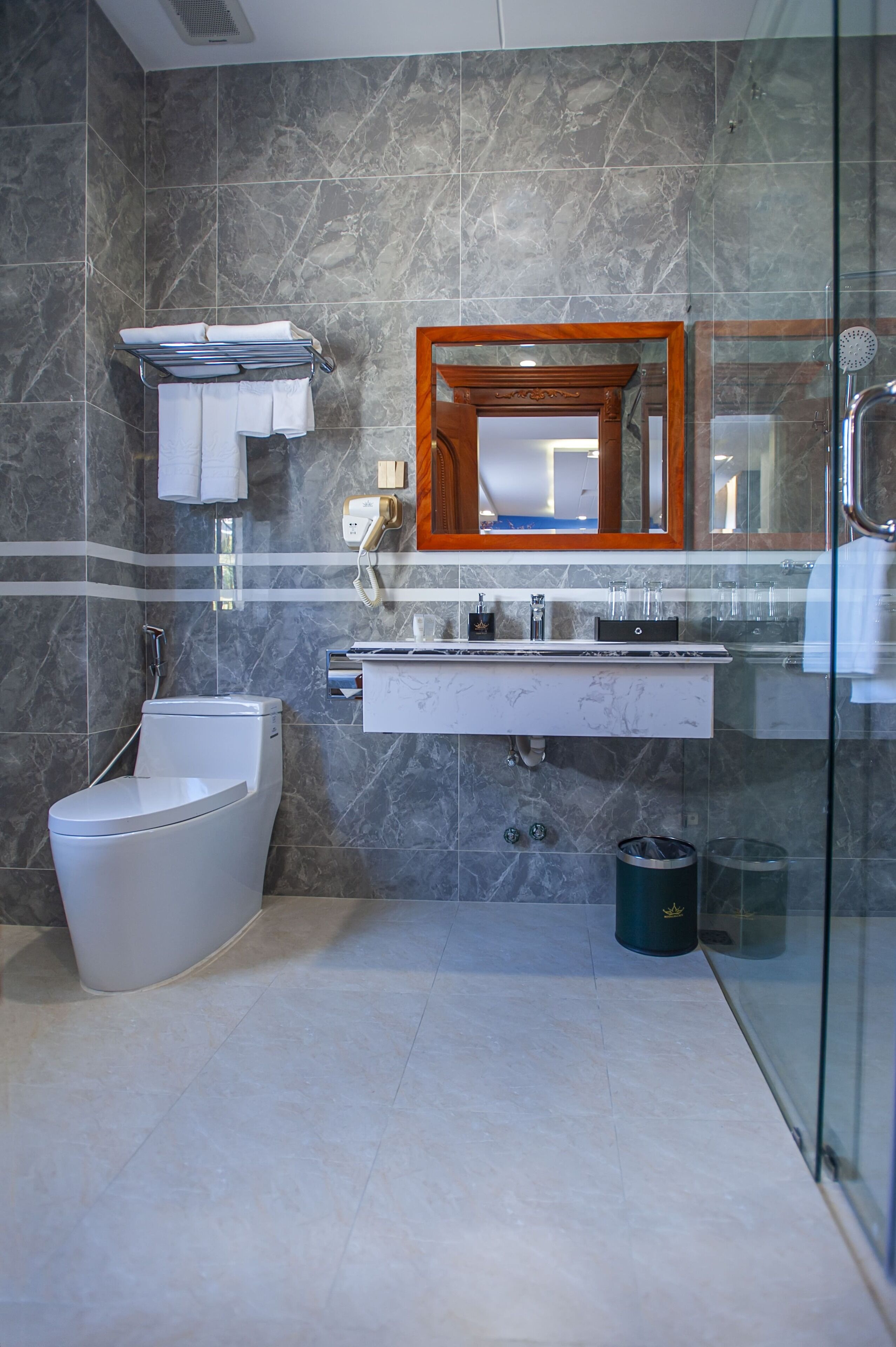 A modern bathroom with a glass shower enclosure, a white toilet, a marble-tiled wall, a wooden-framed mirror, and a glass sink with a marble countertop.