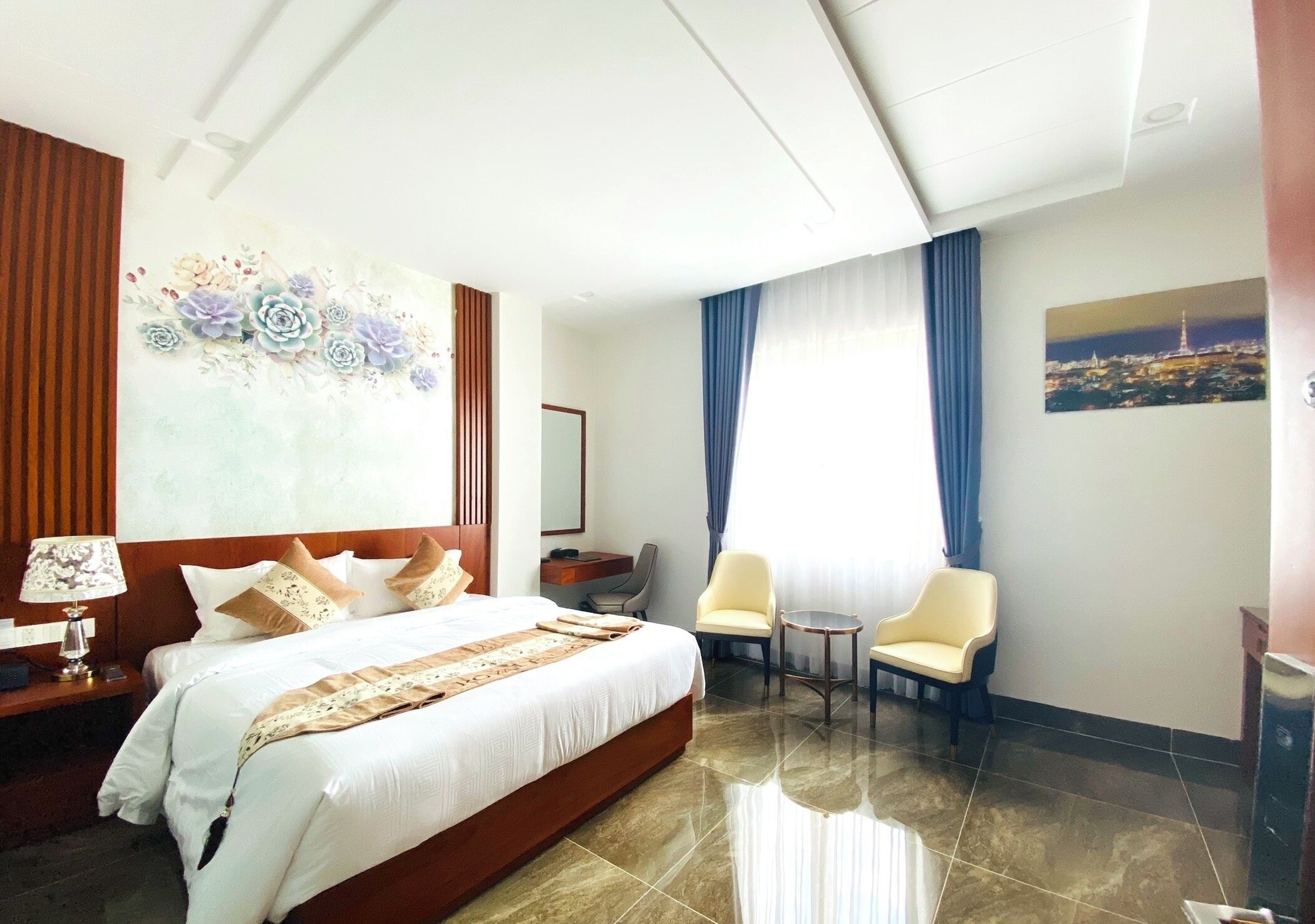 A hotel room with a large bed, a desk with a lamp, two chairs, a small table, and a painting on the wall.