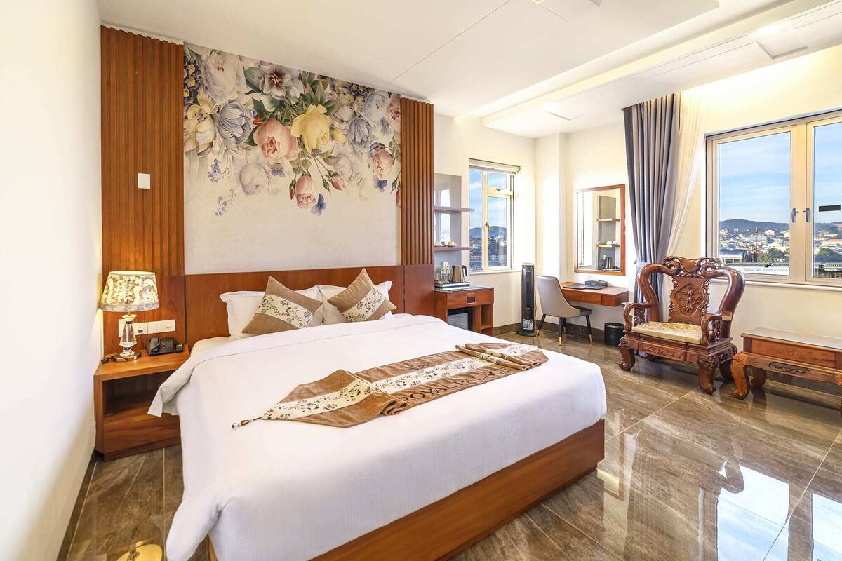 A hotel room with a large bed, a desk, a chair, and a view of the city through the window.