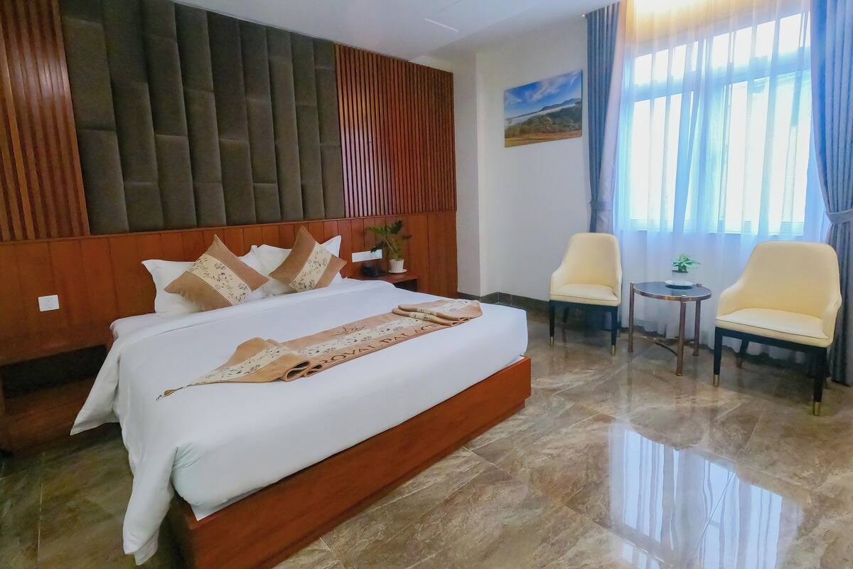 A hotel room with a large bed, a wooden headboard, two armchairs, a small table, and a painting on the wall.