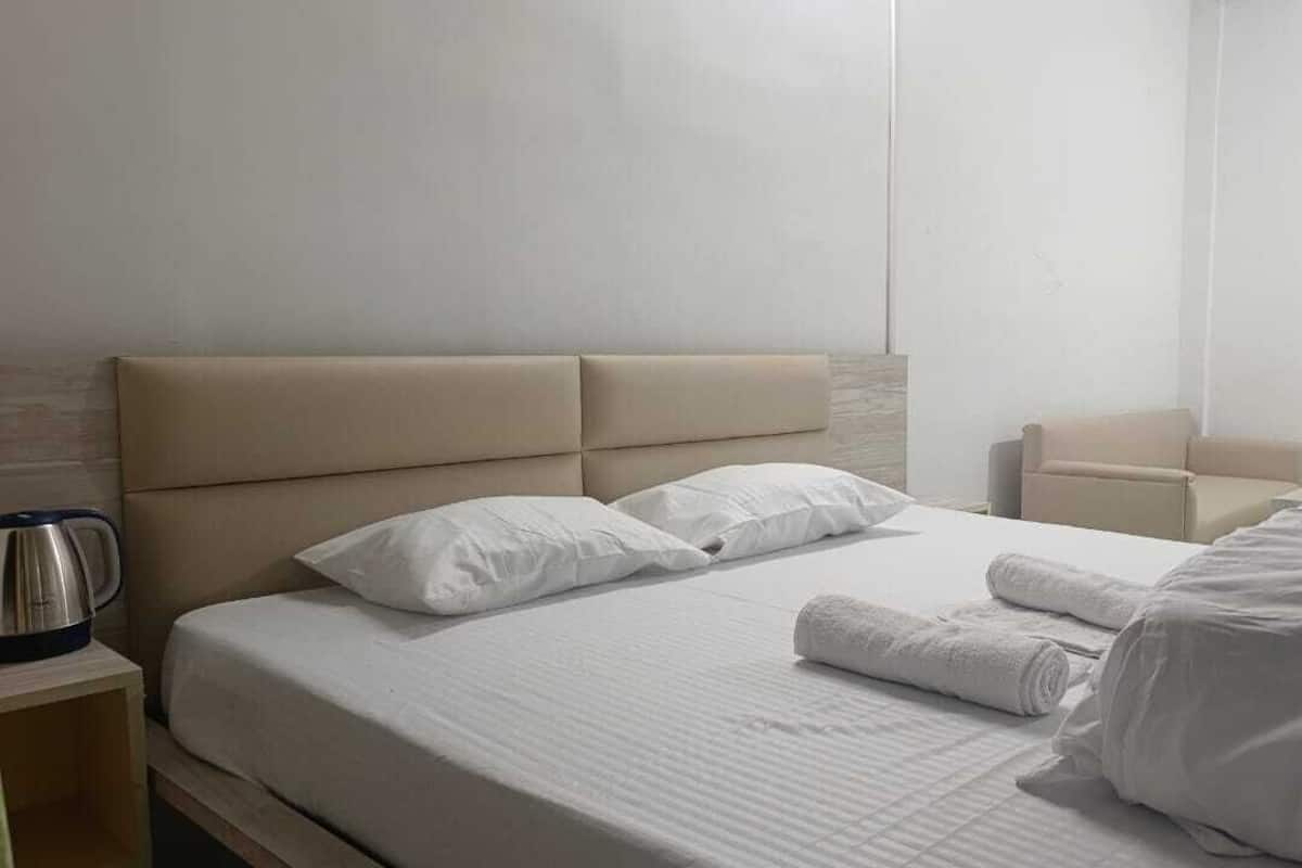 A neatly made bed with white linens, a headboard, and a bedside table with a kettle.