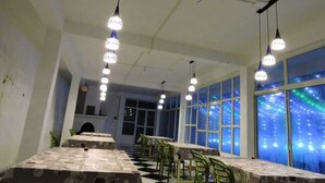 A dining area with long tables and chairs, hanging pendant lights, and large windows with a view of the city at night. - Vinayaka Inn Sittong (Kurseong)